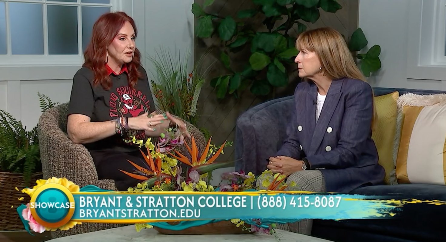 Two women sit and talk on a set adorned with plants and flowers. A banner at the bottom displays “Bryant & Stratton College” along with a phone number and website, encouraging viewers to enroll for the 2026 Fall Semester. The left woman gestures while speaking; the right woman listens attentively.