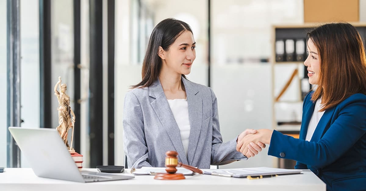 Two women in business attire shaking hands across a desk with legal documents, a laptop, and a gavel on it.