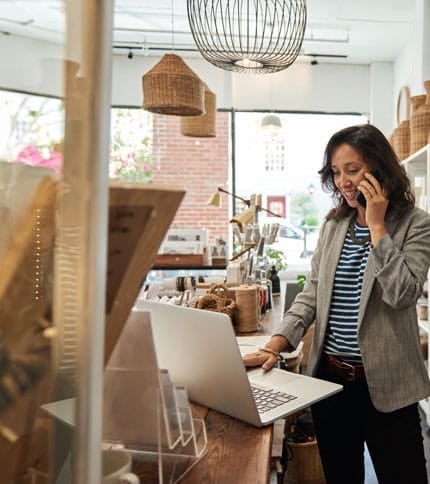 A woman in a striped shirt and blazer stands at a wooden table, talking on her phone while using a laptop. She is in a well-lit shop adorned with wicker baskets and hanging lights.