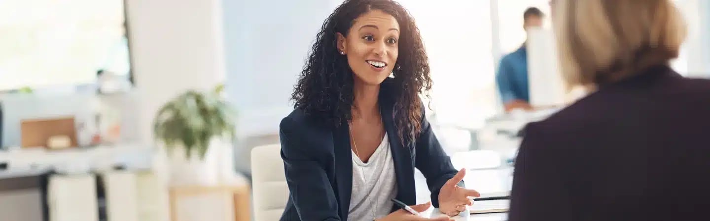 A woman with curly hair talks to a colleague in an office setting, gesturing with her hands.