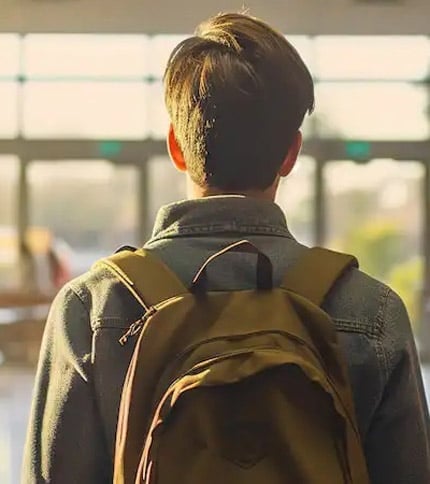 A person with short hair wearing a denim jacket and a brown backpack stands in a brightly lit room, facing large windows that let in sunlight.