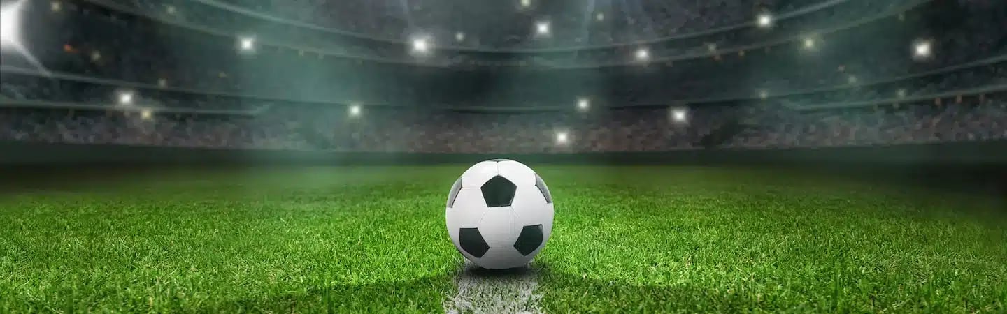 A soccer ball on a green field under bright stadium lights with a blurred crowd in the background.