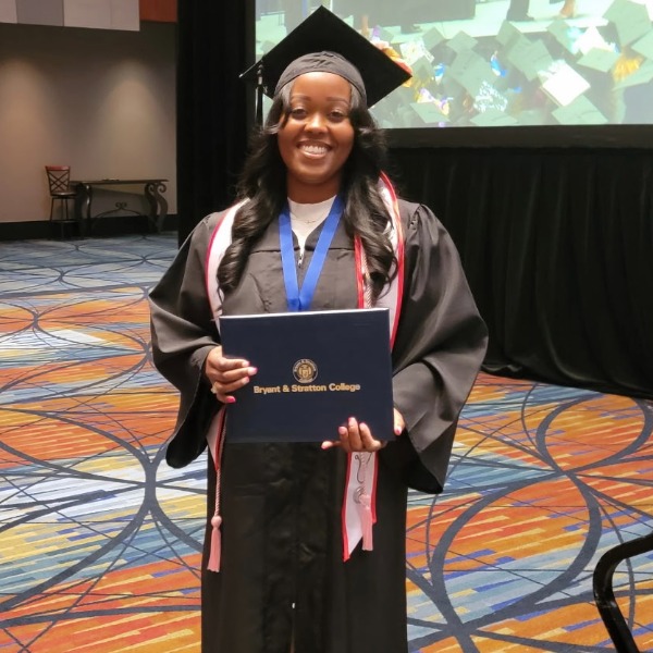 A graduate in cap and gown stands indoors, smiling and holding a Bryant & Stratton College diploma. A projection of graduation caps is visible in the background, celebrating Krystal Whitaker’s accomplishment as an AAS Nursing graduate from Richmond.