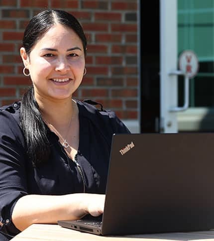 A woman with long dark hair is smiling while sitting outside at a table and working on a ThinkPad laptop. She is wearing a dark blouse and a necklace, and a brick wall with a glass door is in the background.