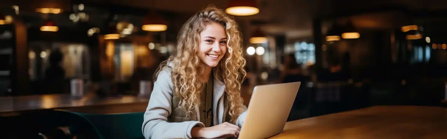 A smiling person with curly hair using a laptop in a warmly lit café.