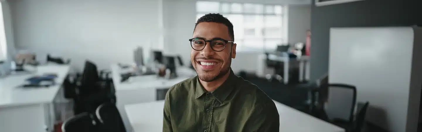 Smiling man wearing glasses and a green shirt in a modern office setting with desks and computers in the background.