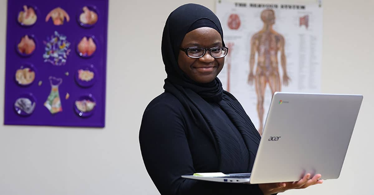 A person in a hijab smiles while holding an Acer laptop in a room with anatomical posters on the wall.