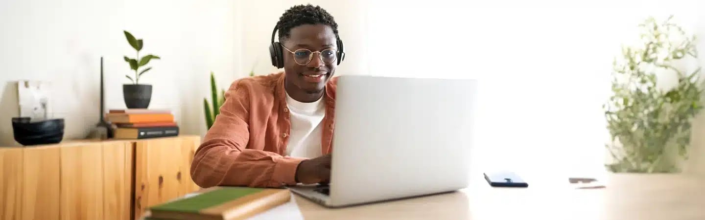 Person smiling and working on a laptop at a desk, wearing headphones. There's a phone, notebook, and bookshelf nearby.