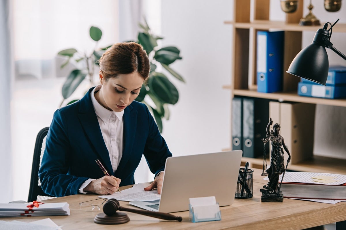 A woman in a suit sits at a desk, writing on papers beside a laptop, with a gavel and Lady Justice statue nearby.