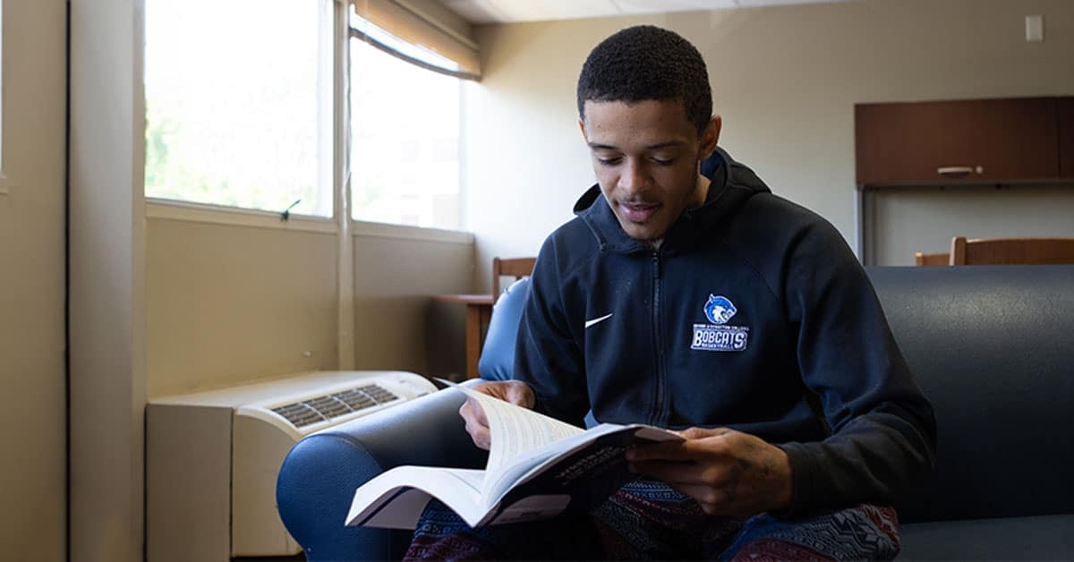 A person sits on a couch reading a book in a well-lit room. The person wears a black hoodie with a logo on it.