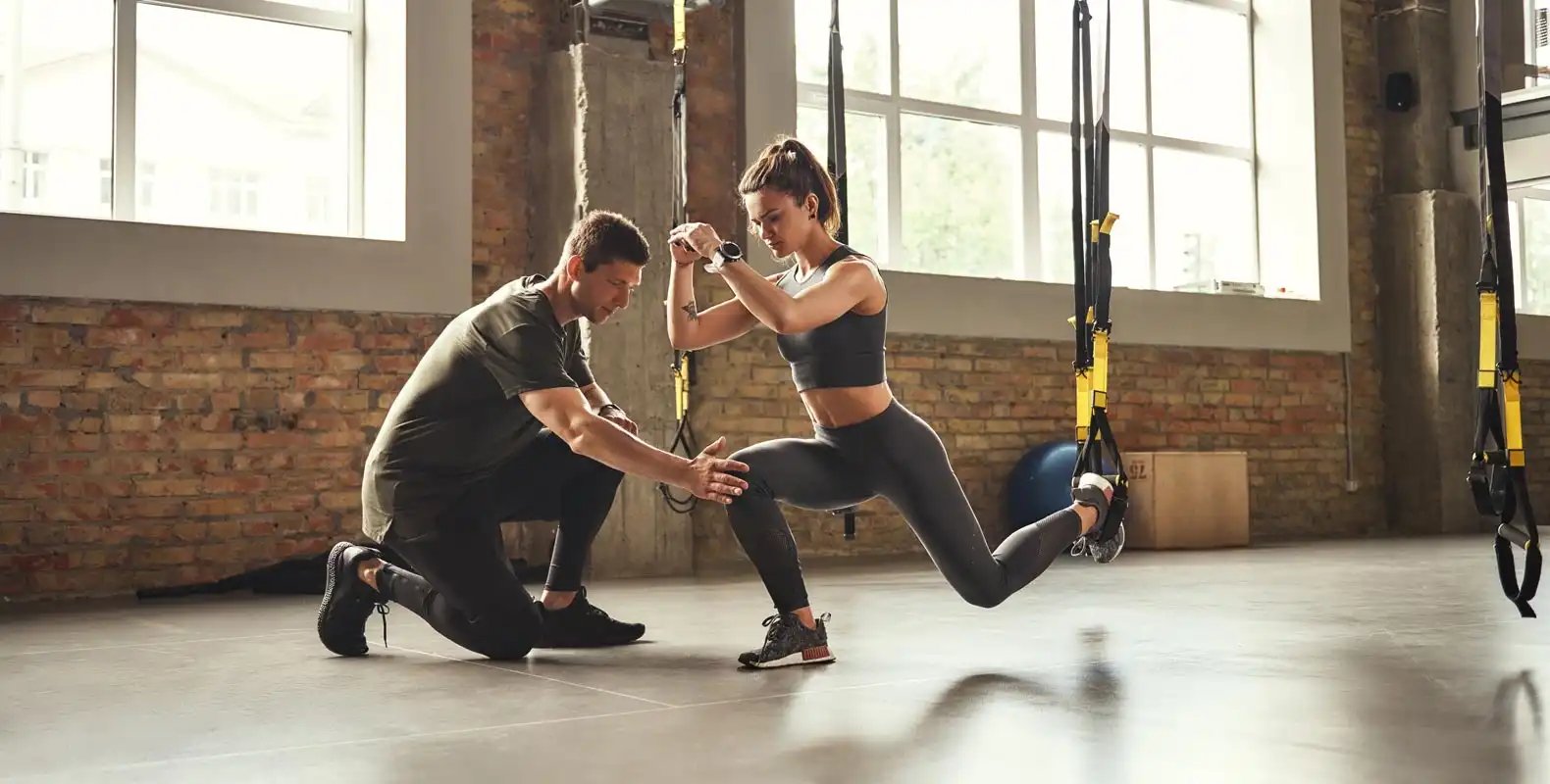 A woman exercises with suspension straps while a man kneels beside her, offering guidance in a gym.