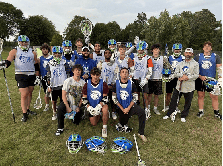 A group of Bryant & Stratton College men’s lacrosse players in blue and white uniforms pose together on a grassy field, most holding sticks and wearing helmets. Trees and a soccer goal are visible in the Wisconsin background.