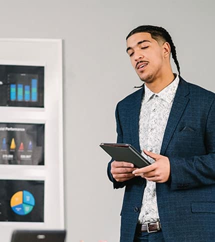 A person in a blue suit stands in an office setting, holding a tablet. Behind them, framed charts and graphs are visible on the wall.