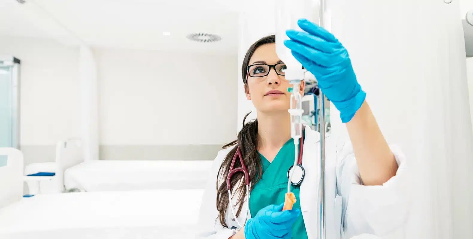 A healthcare professional in a lab coat and blue gloves adjusts an IV drip in a hospital room.