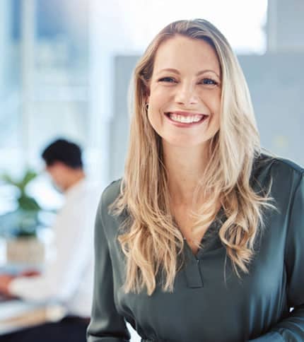 Smiling woman with long blonde hair in a green blouse stands in an office setting. A person is working in the background at a desk, creating a professional atmosphere. The space is bright and modern.