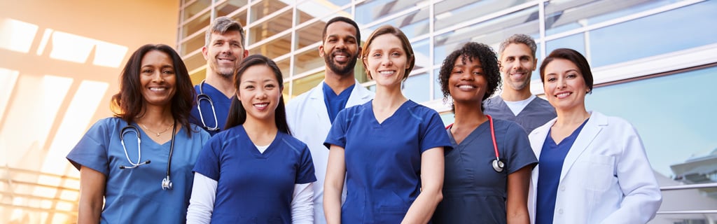 A diverse group of eight smiling healthcare professionals, including doctors and nurses with backgrounds such as an Office Administration degree, stand together outdoors in front of a modern building, wearing scrubs and white coats.
