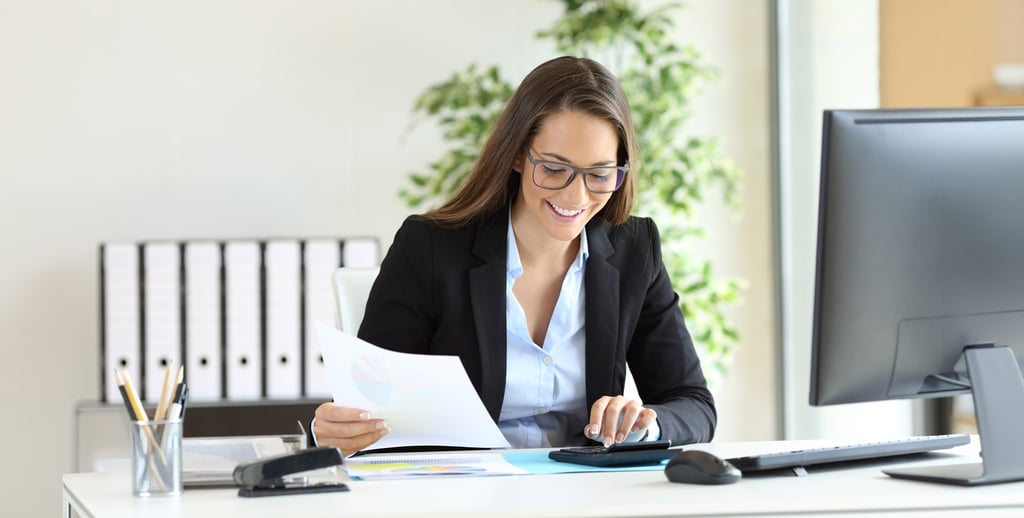 A woman in business attire smiles while using a calculator at her desk, holding papers. Surrounded by office supplies, she reflects the growing demand in accounting careers with shelves and plants in the background.