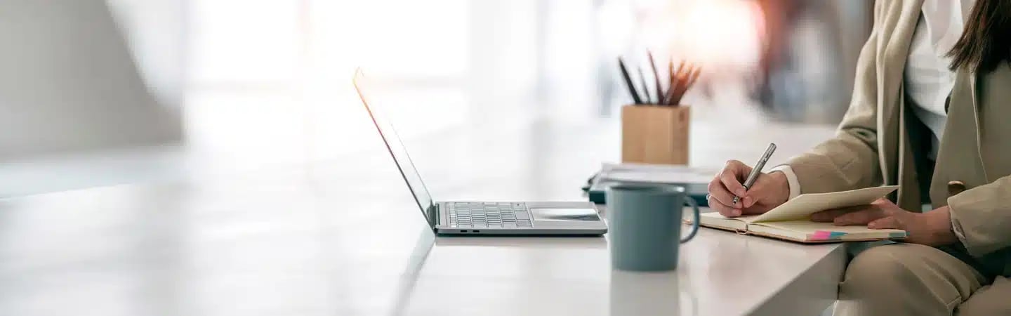 Person in a beige suit writing in a notebook at a desk with a laptop, coffee mug, and pen holder.