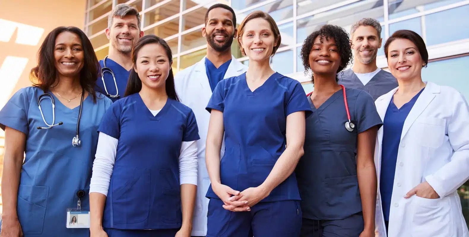 A diverse group of healthcare professionals smiling and posing outdoors in their uniforms.