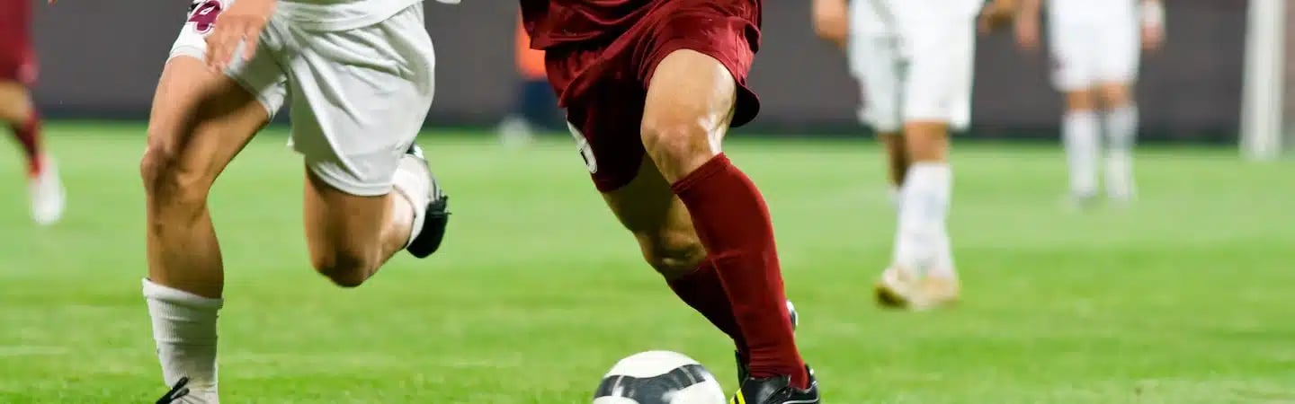 Two soccer players in action, one in white and one in maroon, chasing a soccer ball on a grassy field.