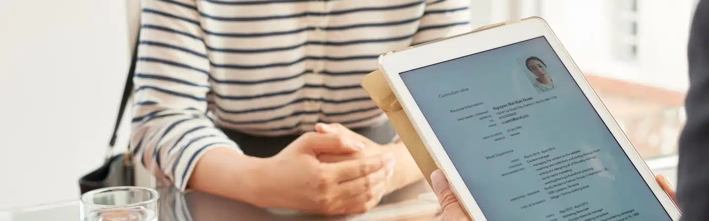 Person holding a tablet showing a resume, while another person sits across the table with hands clasped.