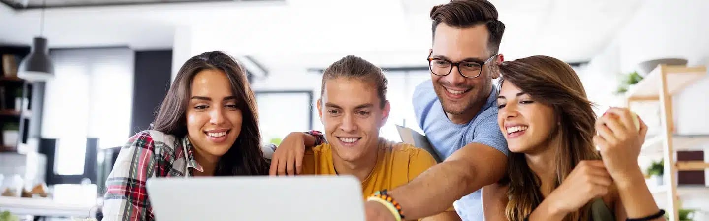 Four people smiling and looking at a laptop screen together in a bright room.