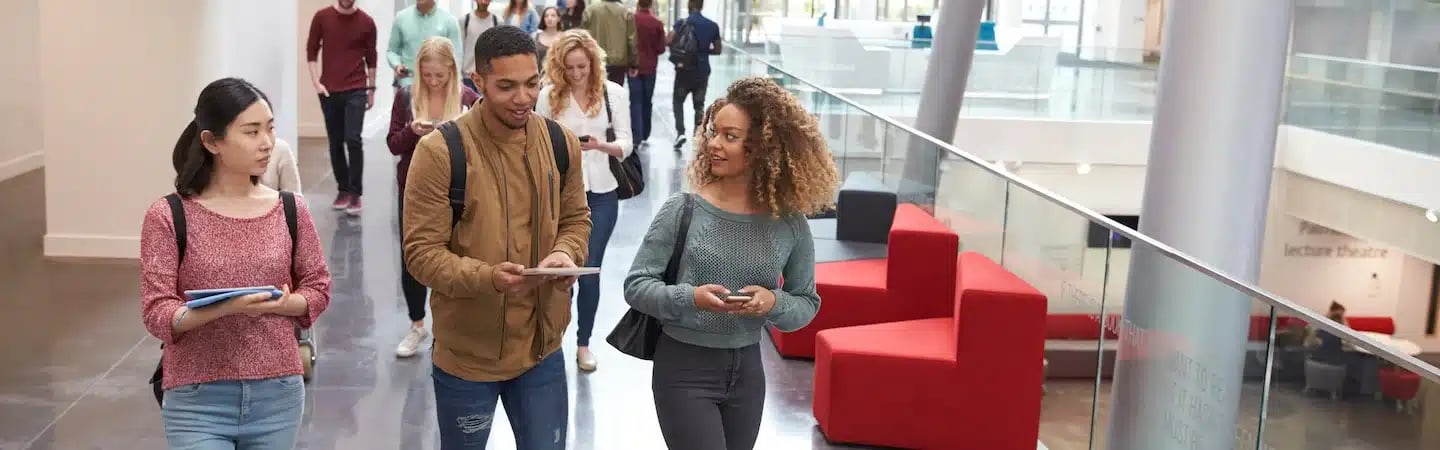 Students walking through a modern university building with glass railings and red seating, engaged in conversation.