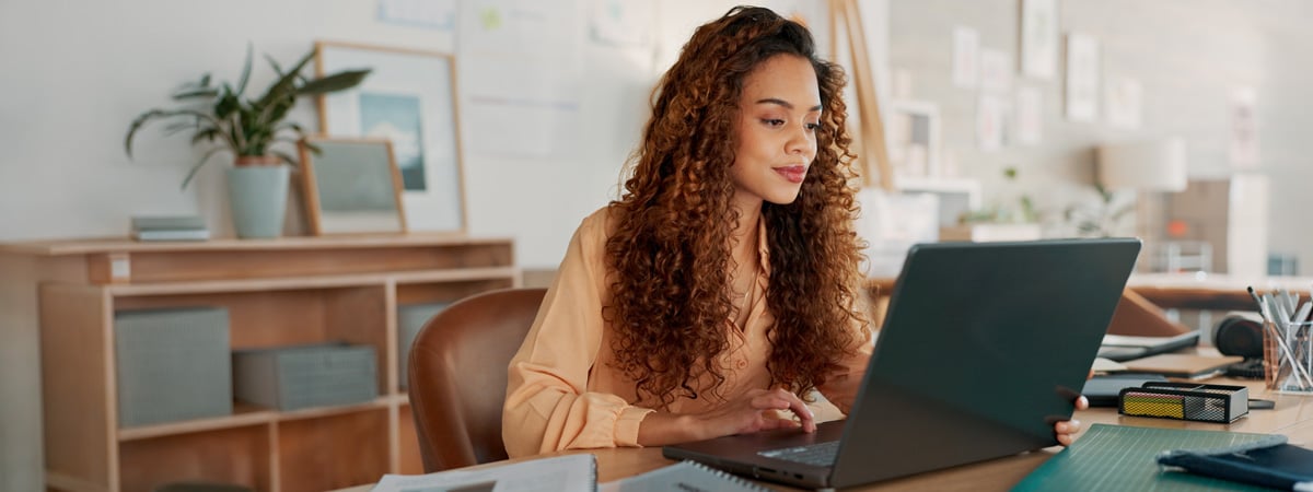 A woman with curly hair sits at a desk in a modern office, looking at a laptop. She appears focused, with papers and office supplies around her, and a plant in the background.