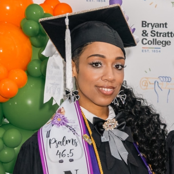 A graduate in cap and gown stands in front of colorful balloons and a Bryant & Stratton College backdrop, wearing decorated sashes—including one celebrating AAS Nursing, and another with “Psalms 46:5” written on it. Chavon Bernard proudly represents Hampton at this special occasion.