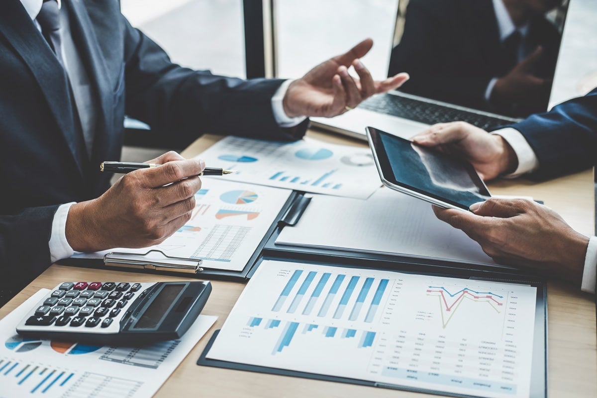 Two people in business suits discussing charts and graphs at a table with documents, a tablet, and a calculator.
