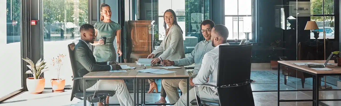 Five people having a meeting around a table in a modern office with large windows and natural light.