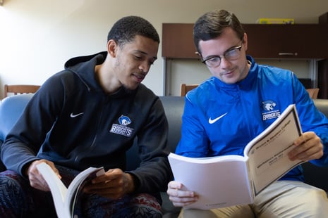 Two people sitting on a couch, studying together and looking at papers. Both are wearing jackets with a bobcat logo.