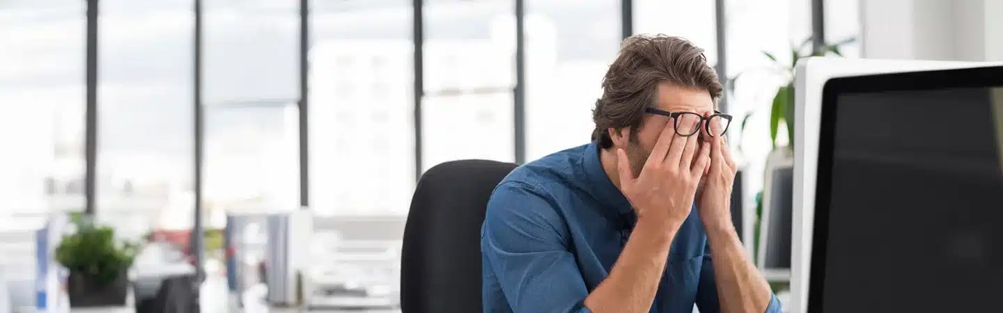 Man sitting at desk in bright office, wearing glasses, and rubbing his eyes while looking at a computer screen.