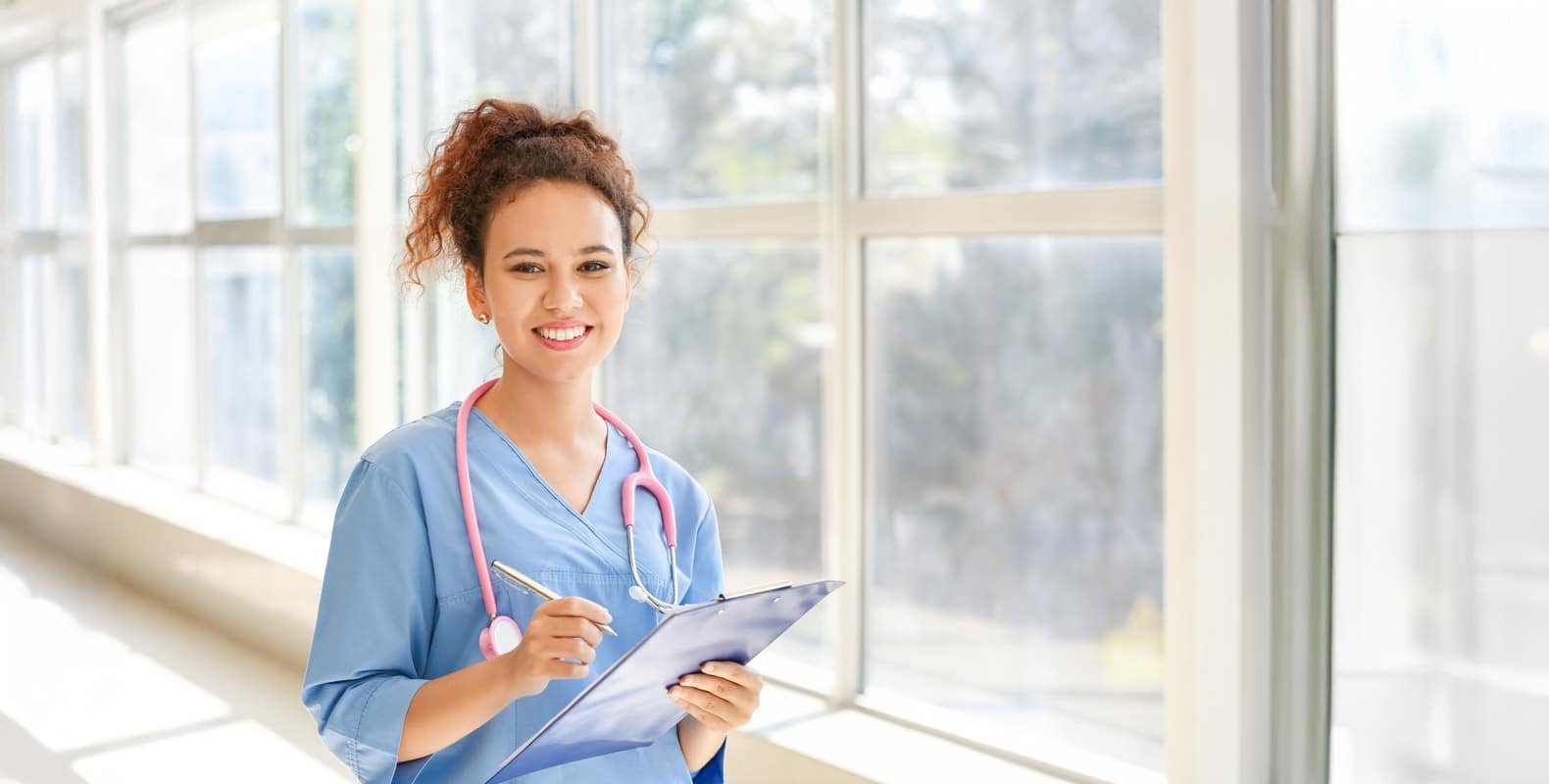 A smiling nurse in blue scrubs holds a clipboard in a bright, sunlit hallway.