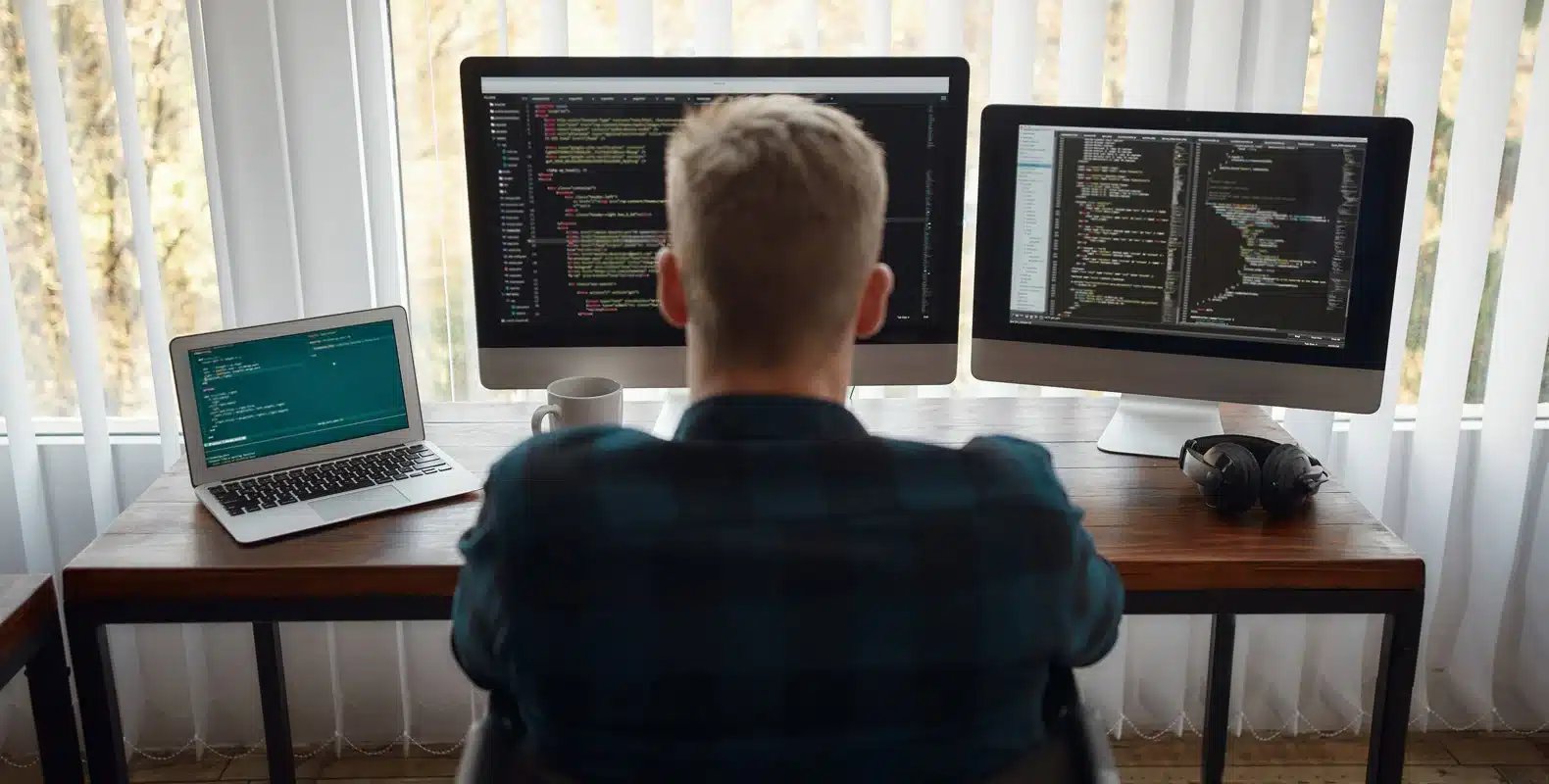 A person sits at a desk with a laptop and two monitors displaying code, in front of a large window with sheer curtains.