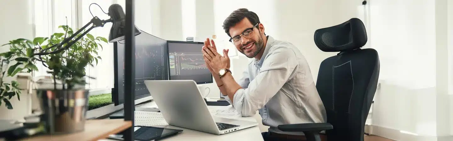 Man with glasses sitting at a desk with multiple monitors, a laptop, and office plants, smiling while clapping his hands.