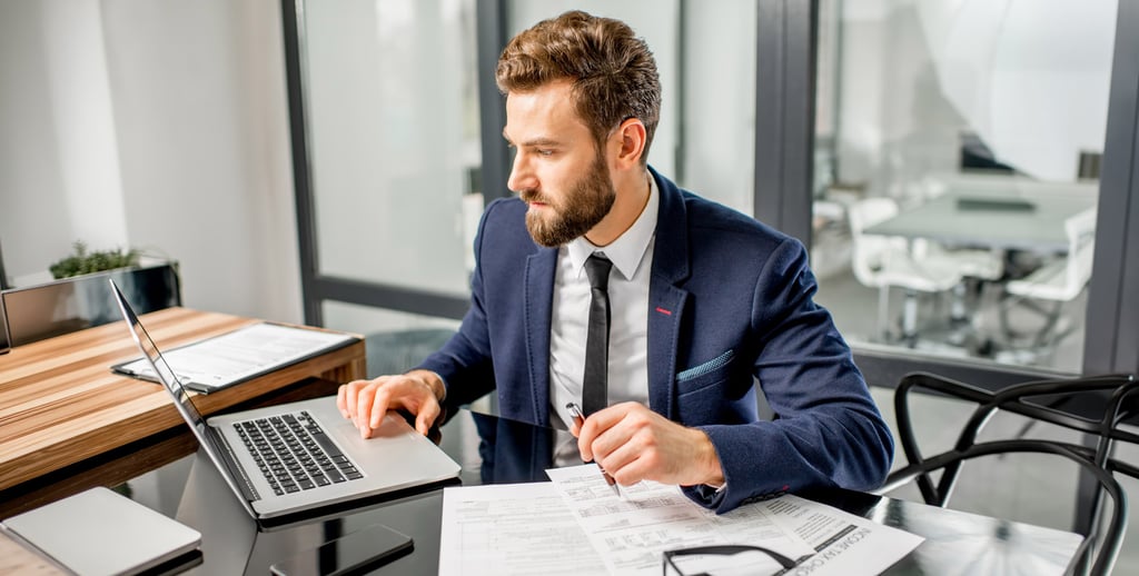 A man in a suit, considering accounting careers, works at a desk with a laptop and paperwork, holding a pen and looking to the side in a modern office setting.