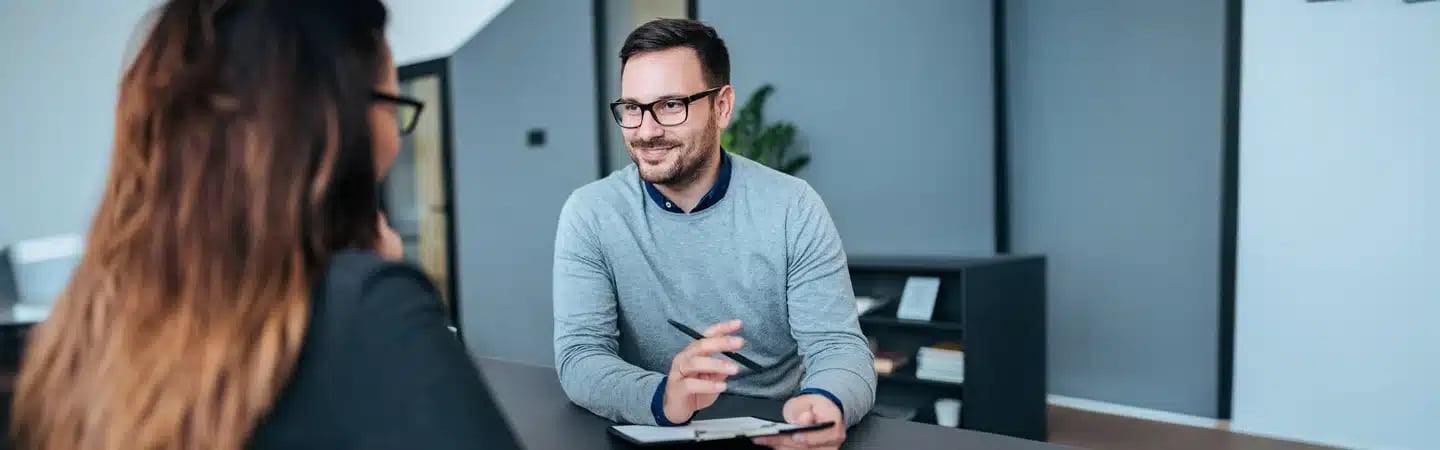 Man in glasses and sweater speaking with a woman across a desk in a modern office setting.