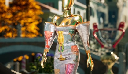 Mannequin with a metallic finish wearing a colorful embroidered dress on a street with buildings and a canal in the background.
