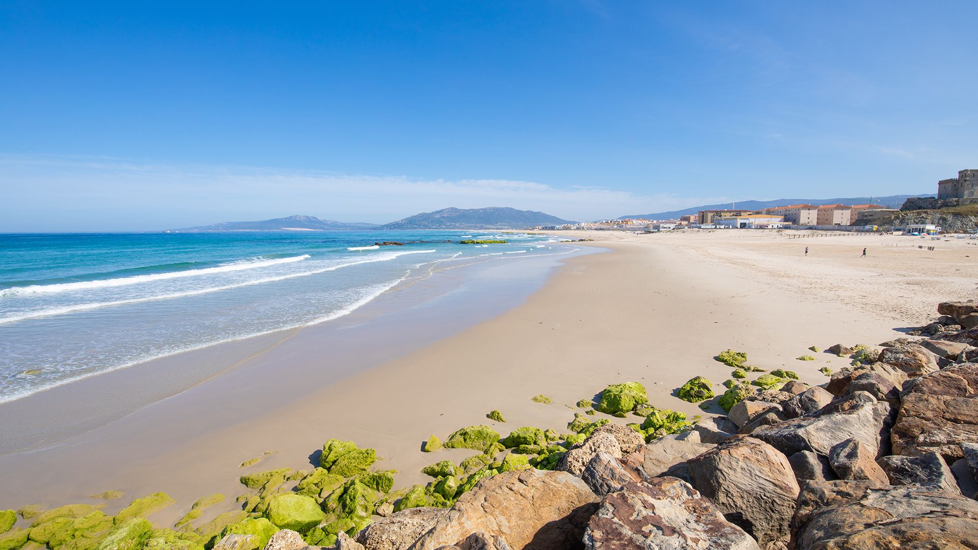 Balneario Kitesurfing in Tarifa with the shoreline, stones in front and a beautiful endless beach with sunny blue sky and mountains in the background
