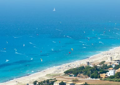 Lefkada Beach Kitesurfing in Greece aerial view on the white sand and bright blue ocean with about 50 kitesurfers on the water and one sailing boat in the background