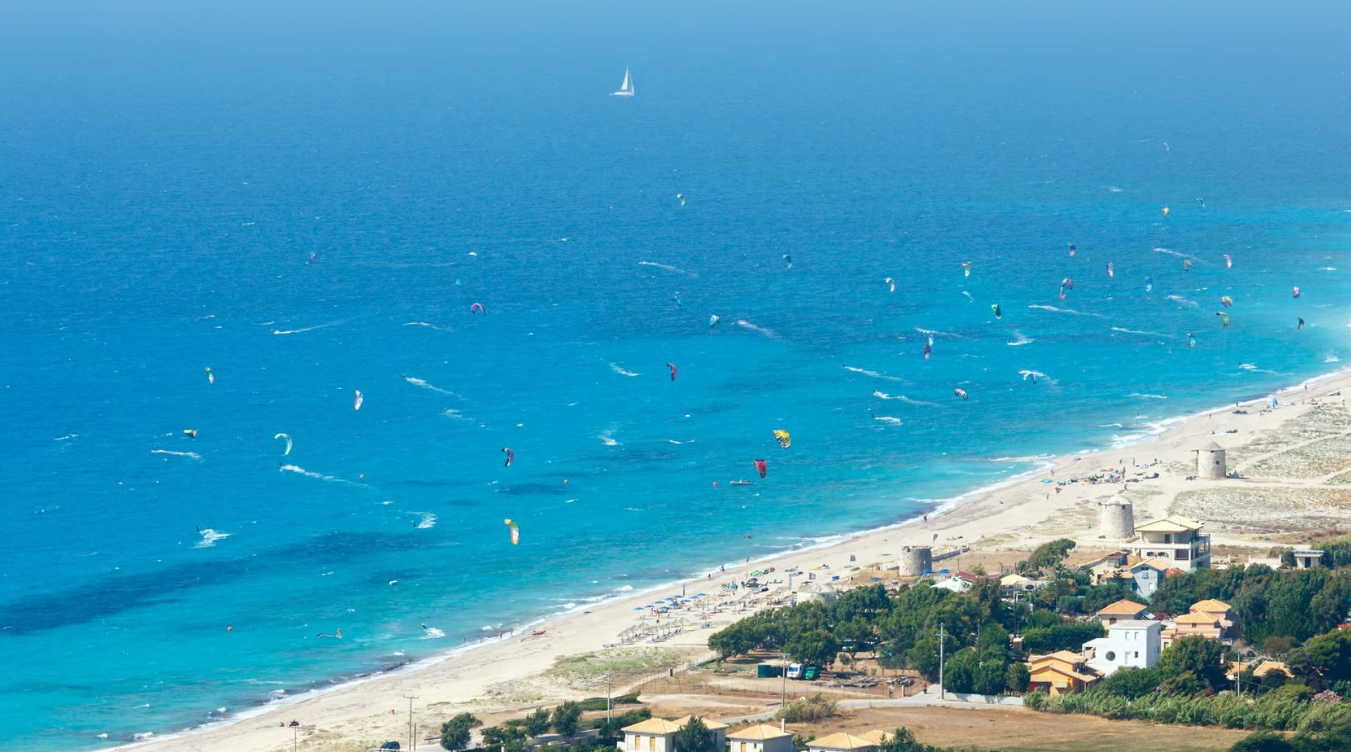 Lefkada Beach Kitesurfing in Greece aerial view on the white sand and bright blue ocean with about 50 kitesurfers on the water and one sailing boat in the background