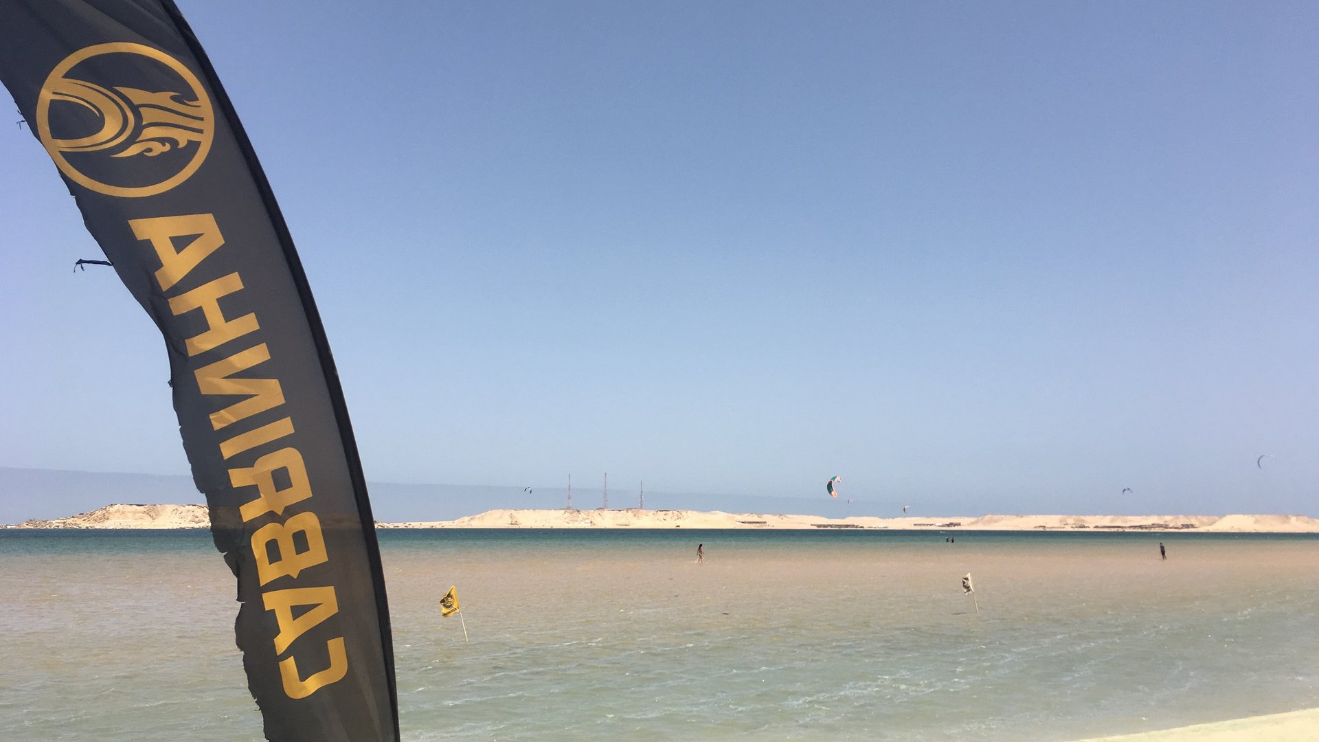 Dakhla Speed Spot Kitesurfing flag in front with Kitesurfers, sea and dunes in the background