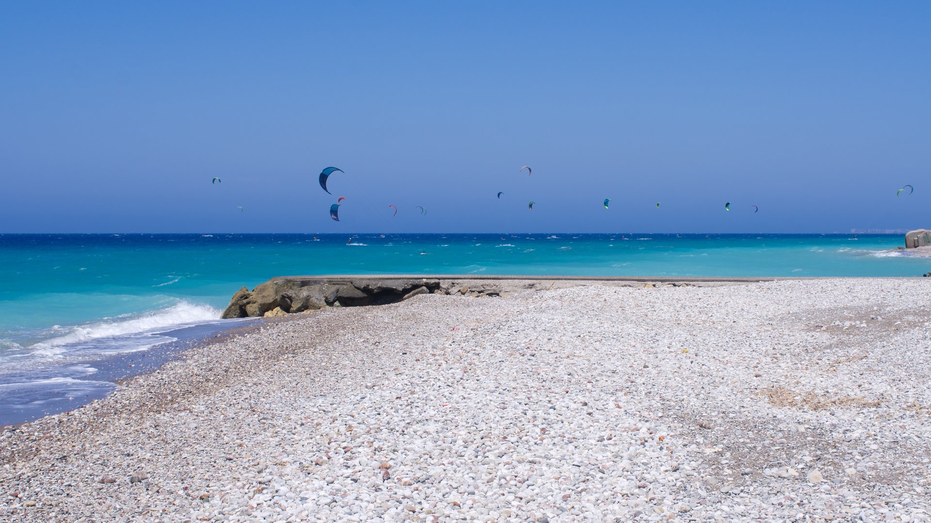 Kremaski Beach Kitesurfing in Rhodos stony Beach with a cliff and kitesurfers on the water in the background on a blue sky sunny day