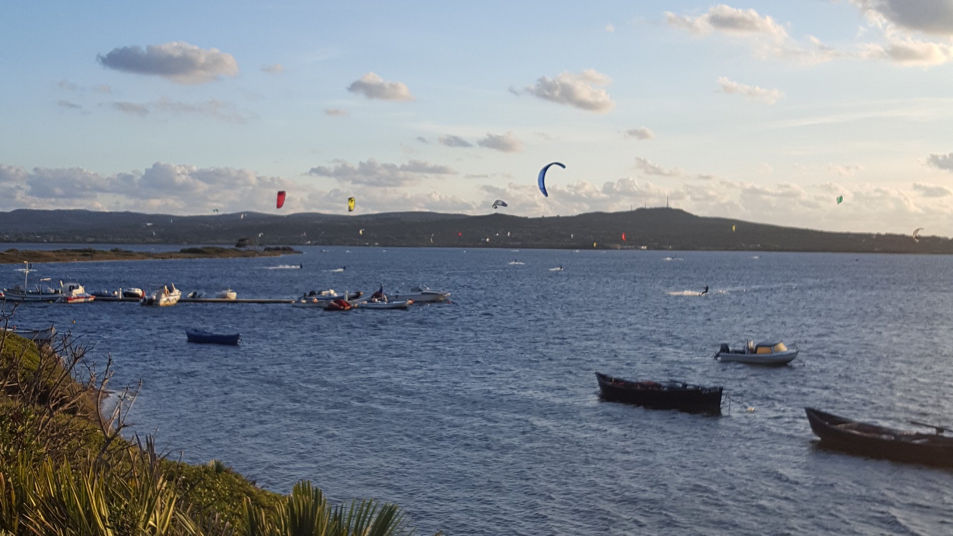 Punta Trettu Kitesurfing in Italy view from the land to the sea with about 6 Kites in the sky and some small fisher boats in front