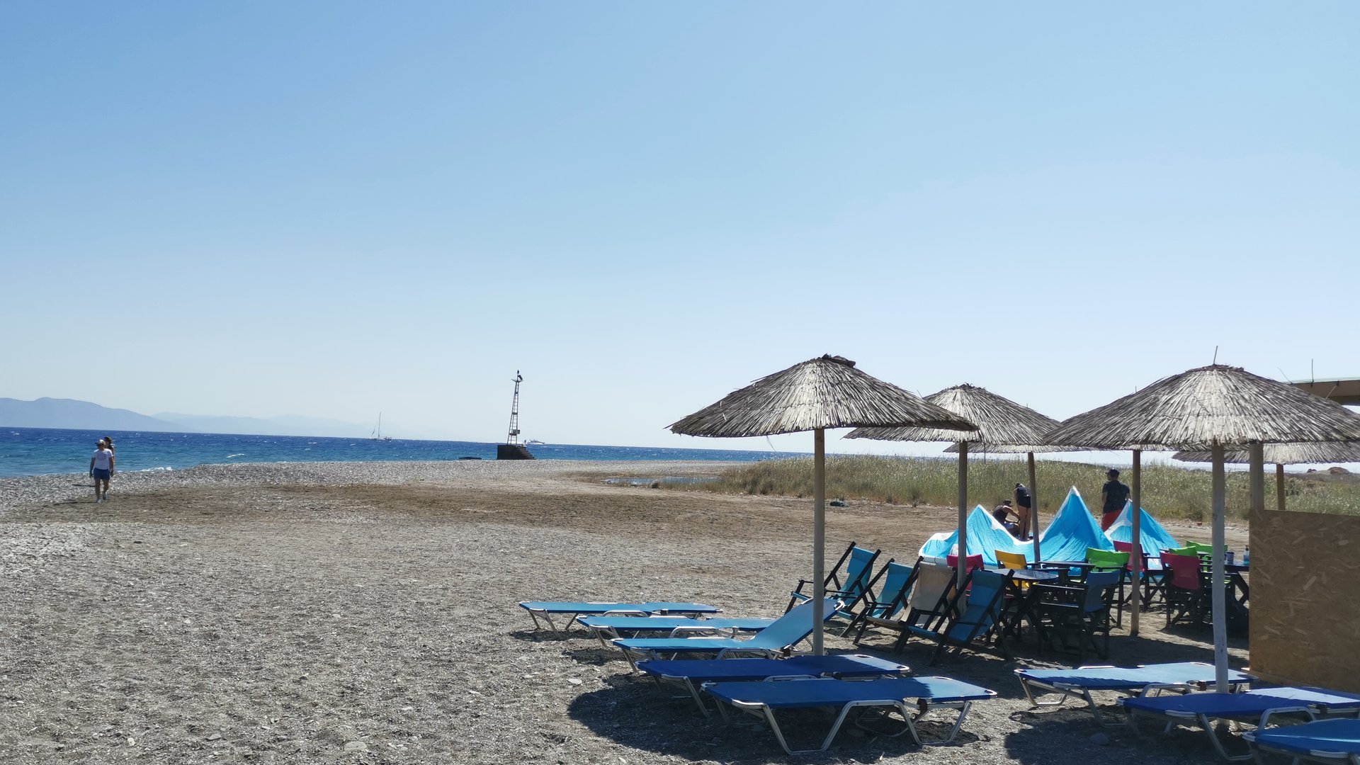 Psalidi Beach Kitesurfing in Kos stony beach with some blue lounge chairs and straw sunshades