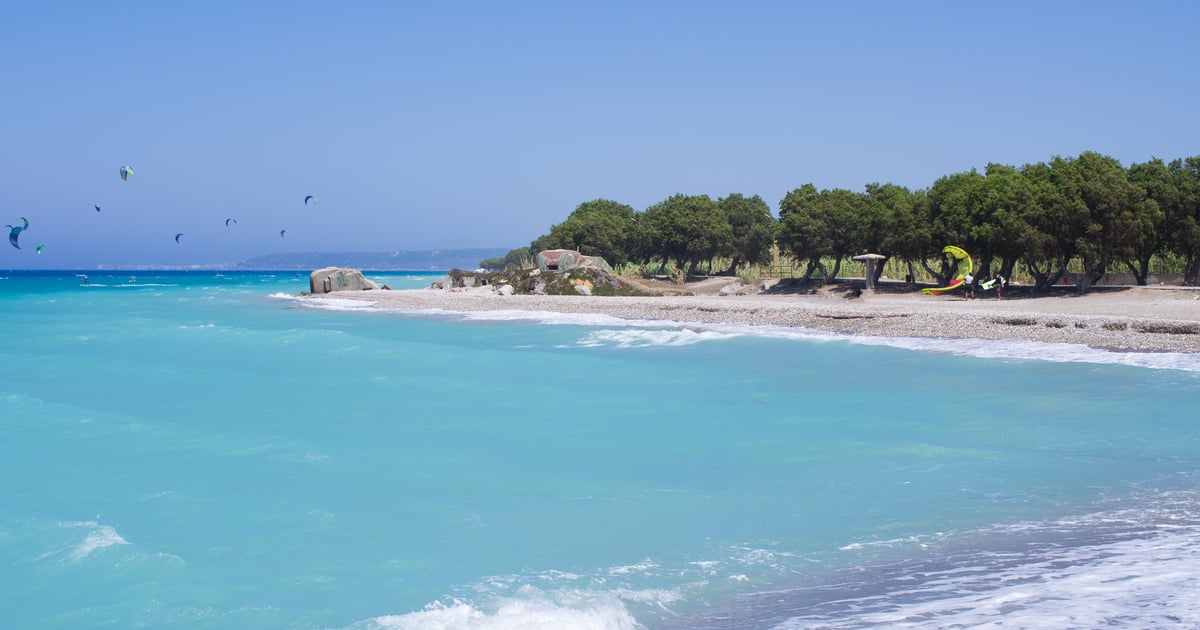 Kremaski Beach Kitesurfing in Rhodos stony Beach with a cliff and kitesurfers on the water in the background on a blue sky sunny day