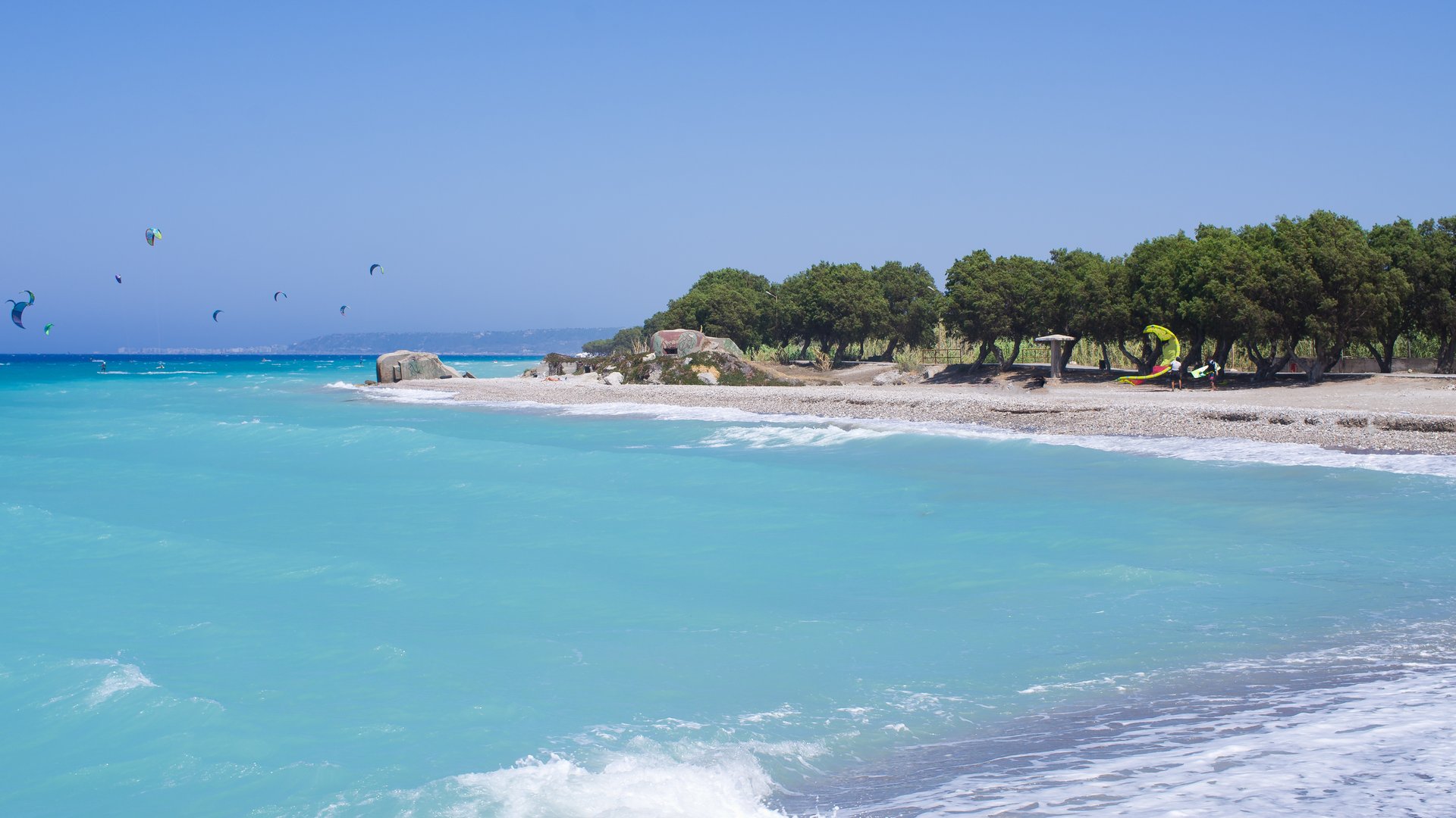 Kremaski Beach Kiteboarding in Rhodos Greec with bright blue water green trees and kiters in the background surfing on the water