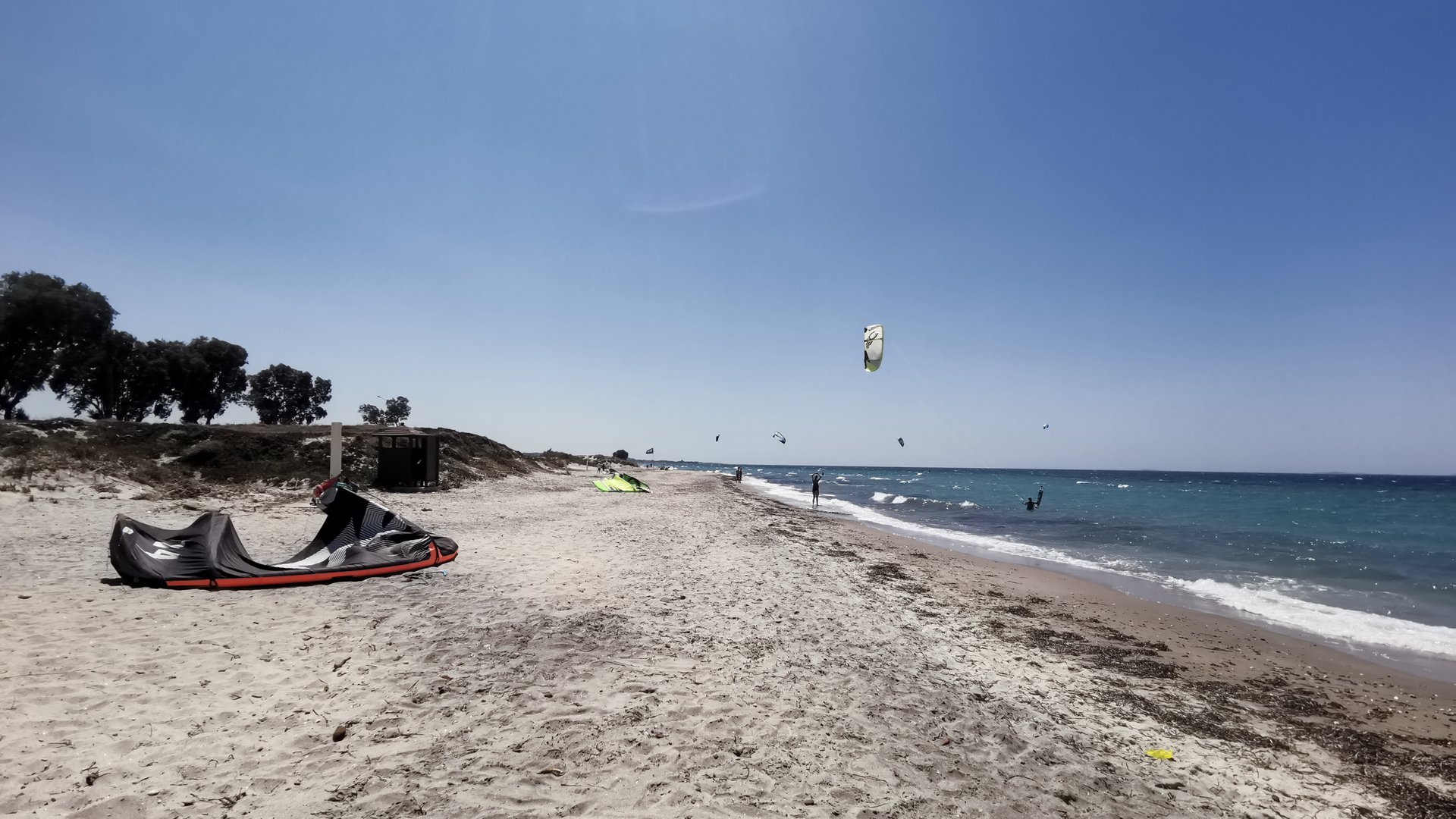 Marmari Beach Kitesurfing in Kos picture from the white sandy beach with a kite laying in front and some trees and surfers in the background