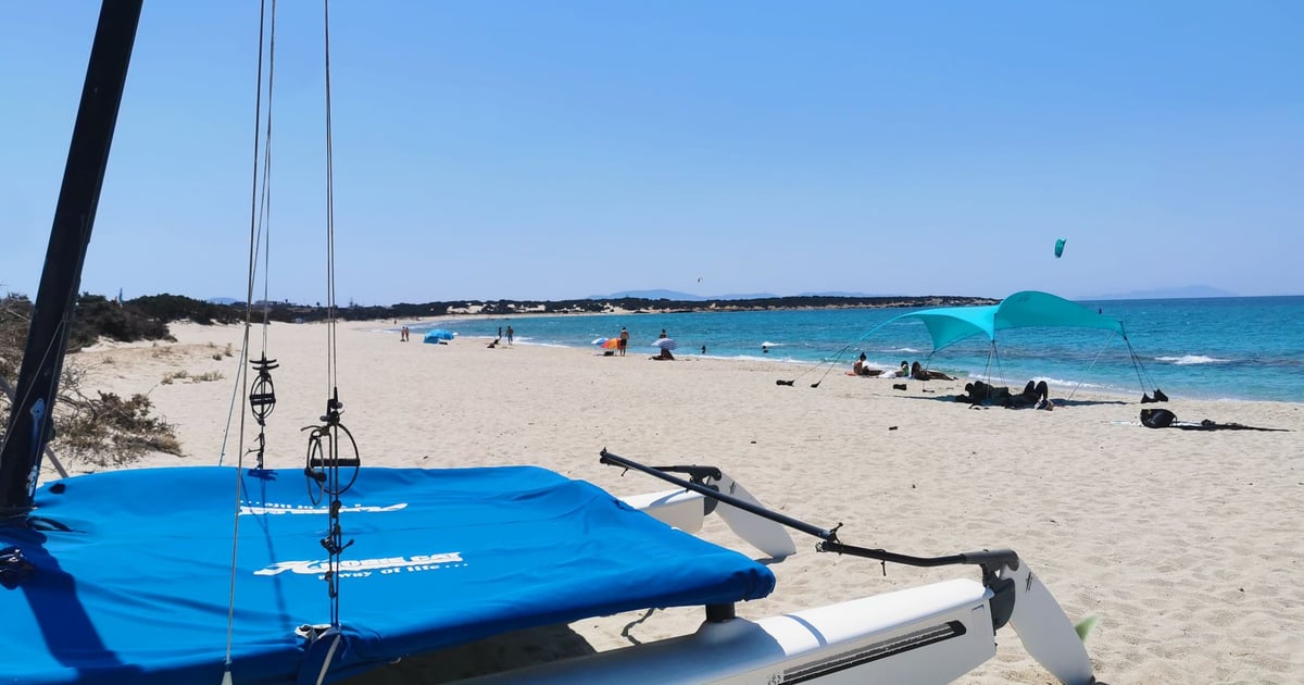 Paralia Glifada Kitesurfing in Naxos Greece with a catamaran boat in front on white sand, some people laying on the beach and a few kitesurfers surfing in the background